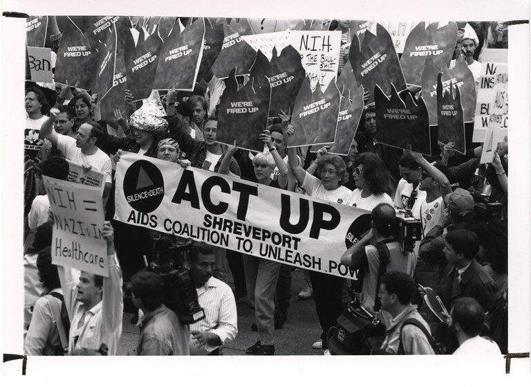 uomini e donne manifestano negli anni 90 in una strada americana con uno striscione su cui è scritto Act Up