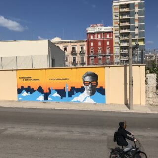 Murales su Piepaolo Pasolini su fondo giallo con scritta "Ti dicono di non splendere e tu spelndi invece". Accanto un ritratto di Pasolini con un paio di occhiali e sullo sfondo lo skyline di Bari.
