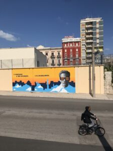 Murales su Piepaolo Pasolini su fondo giallo con scritta "Ti dicono di non splendere e tu spelndi invece". Accanto un ritratto di Pasolini con un paio di occhiali e sullo sfondo lo skyline di Bari.