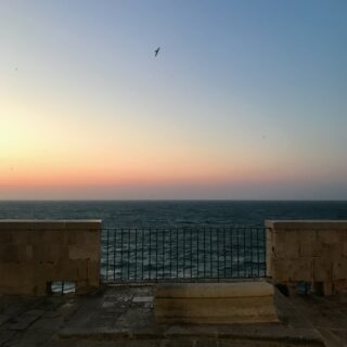 vista da un lungomare al tramonto, il mare sullo sfondo e il muretto e una ringhiera in primo piano