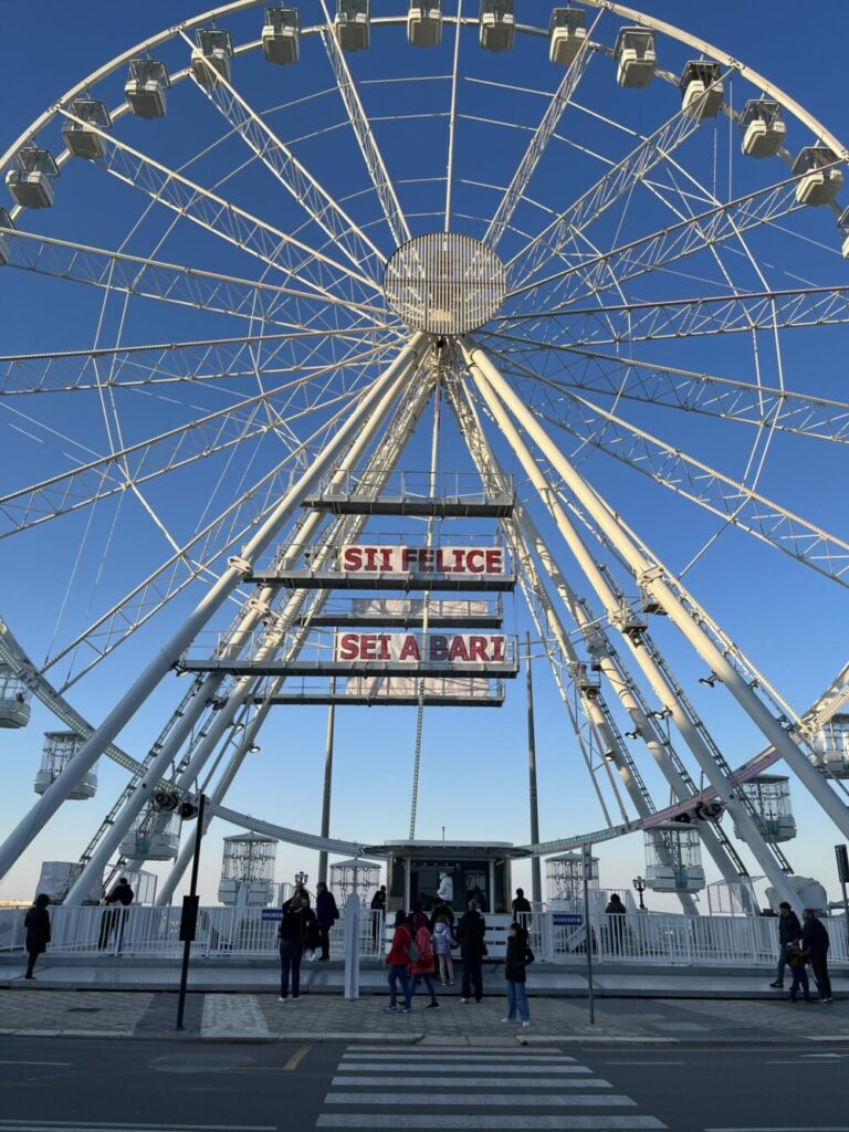 ruota panoramica nel cielo blu del lungomare di Bari. Una scritta rosa al centro: Sii felice sei a bari.